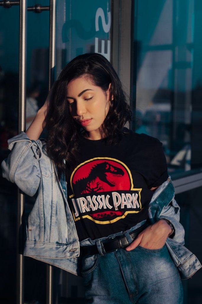 Stylish woman in graphic tee and denim jacket posing against a glass door, bathed in sunlight.